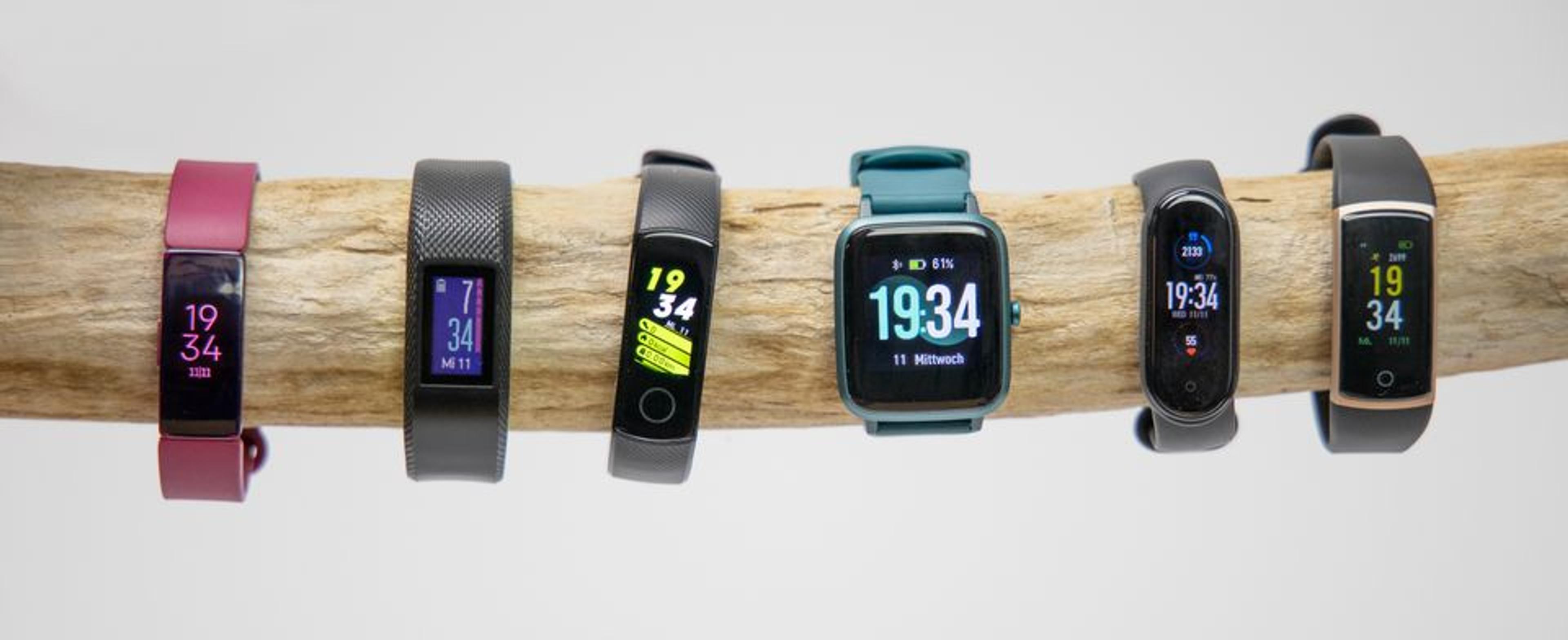 Sieben verschiedene Fitness-Tracker und Smartwatches nebeneinander auf einer Holzstange aufgehängt