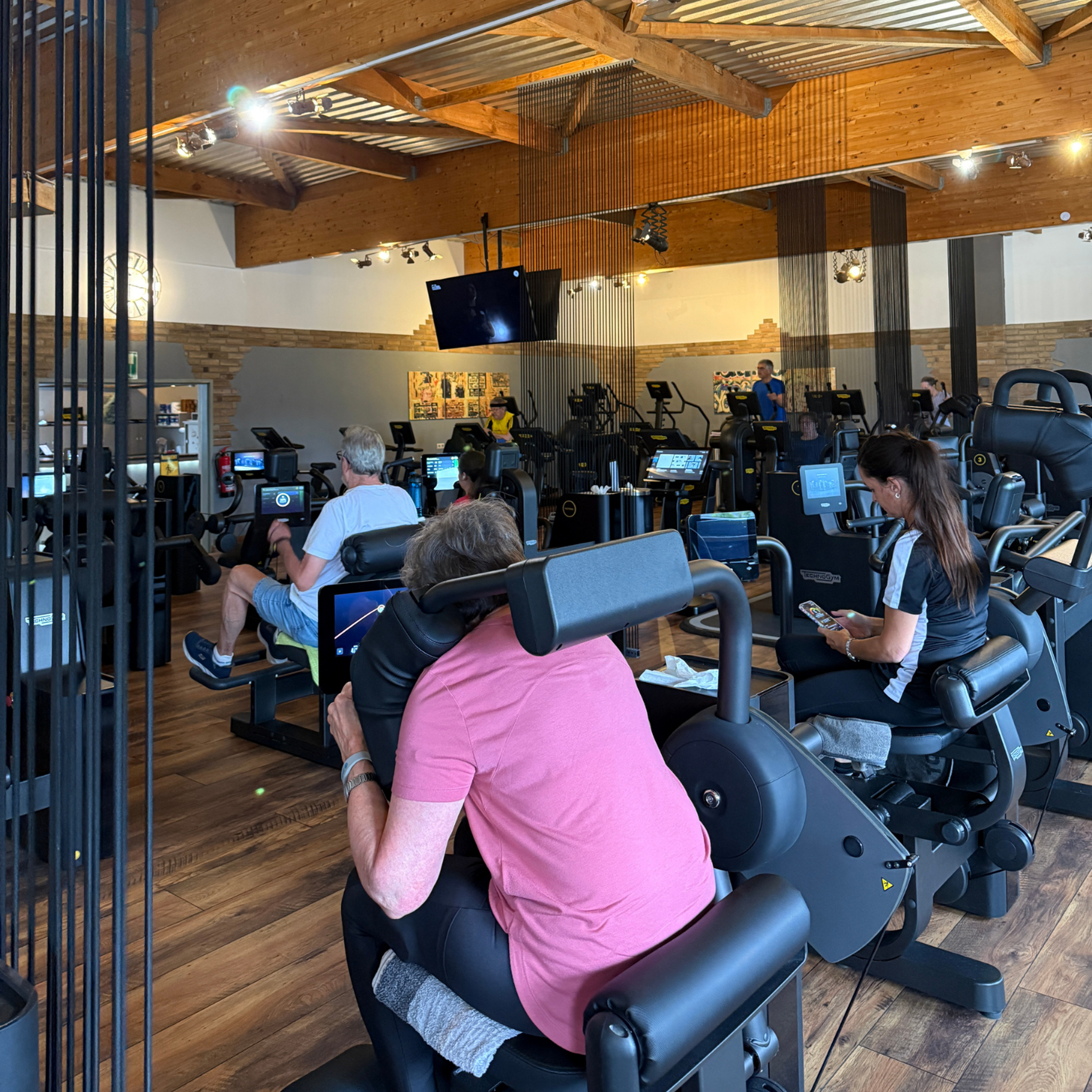 Eine Innenaufnahme eines Fitnessstudios mit mehreren Personen an Sitz-Kraftgeräten mit integrierten Bildschirmen, sichtbaren Holzträgern und Holzfußboden.