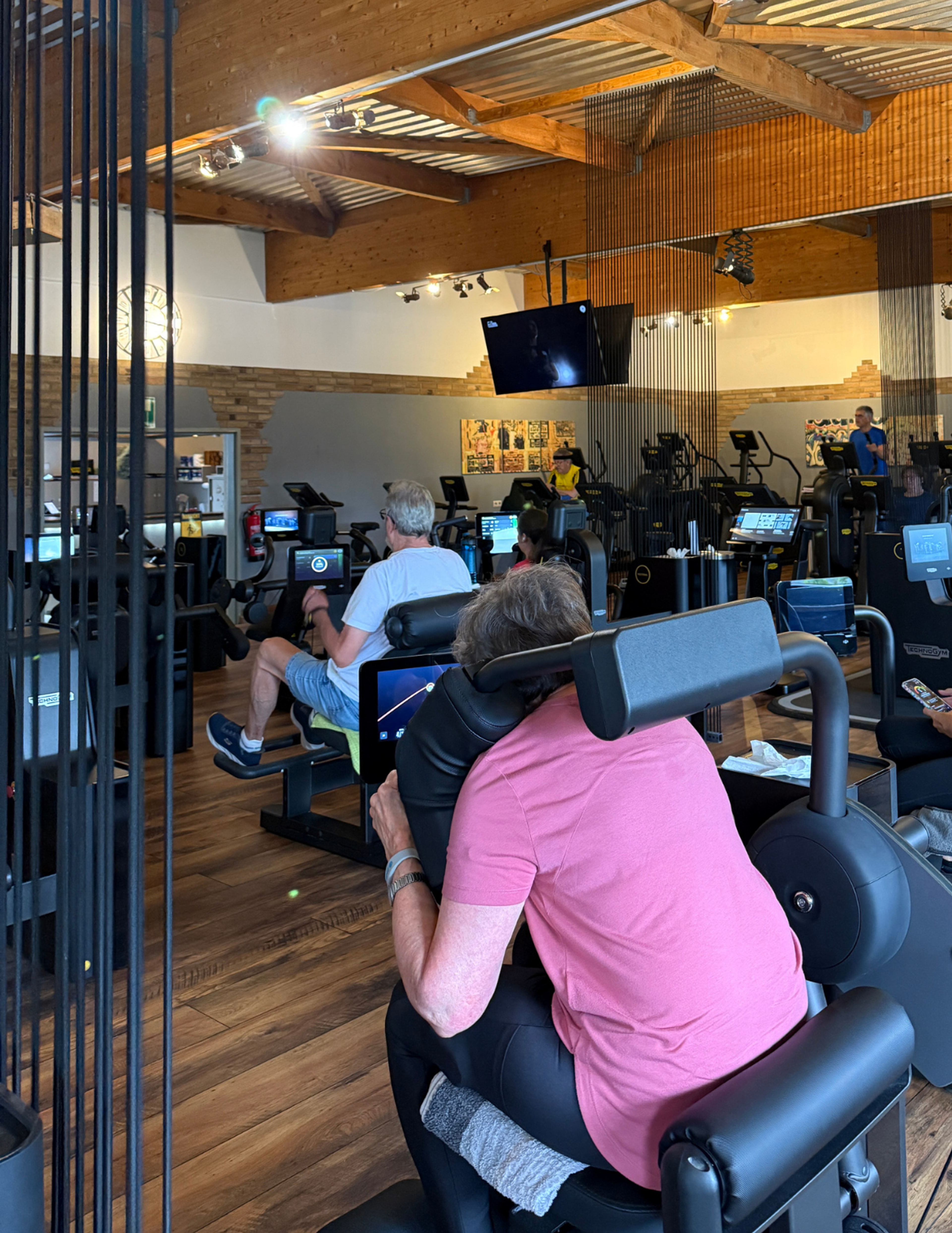 Mehrere Personen trainieren an sitzenden Kraftgeräten in einem hellen Fitnessstudio mit sichtbaren Holzbalken an der Decke, Geräte-Monitoren und einem an der Decke montierten Fernseher.