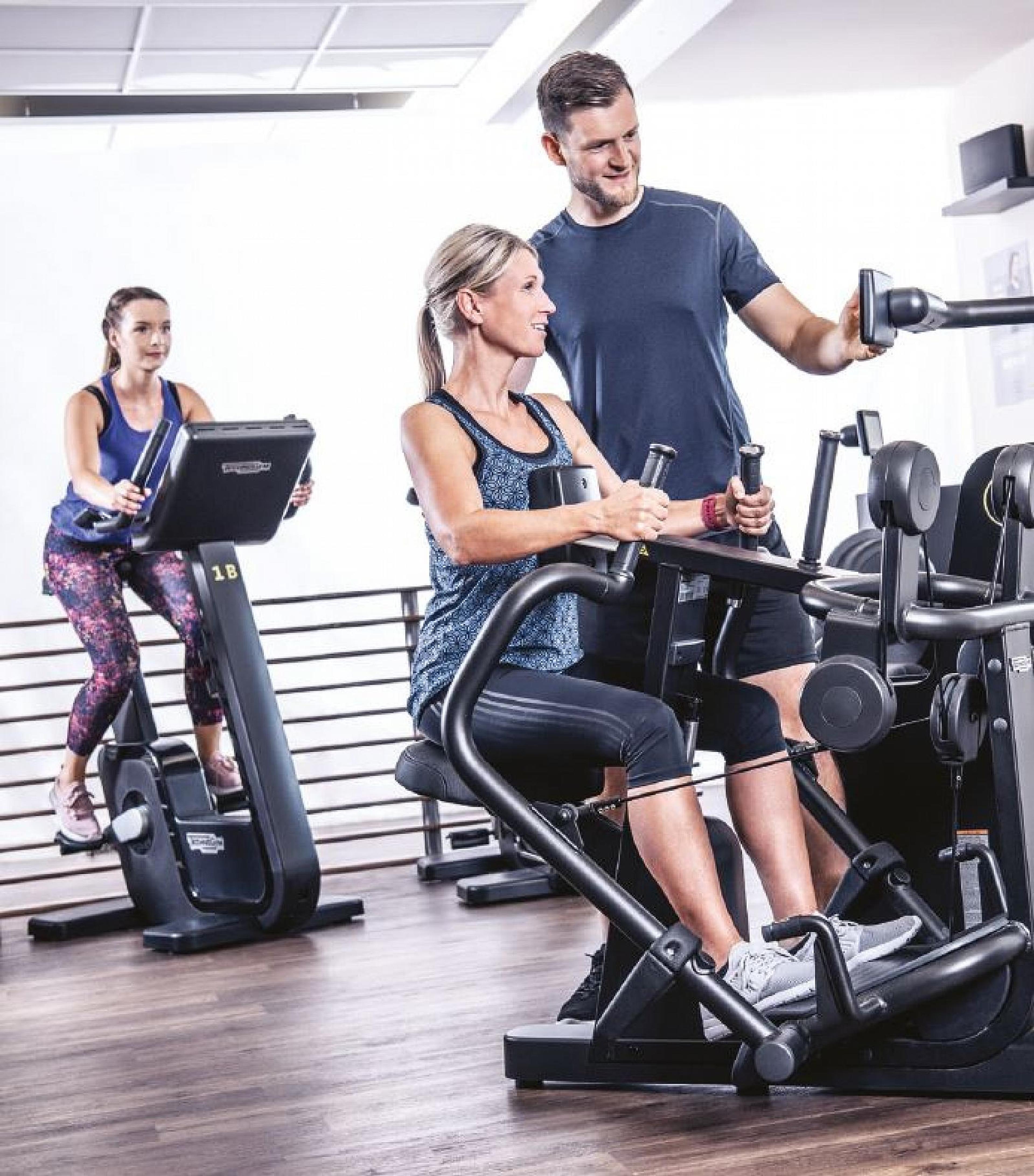 Zwei Frauen trainieren an Fitnessgeräten, eine davon mit Unterstützung eines Trainers