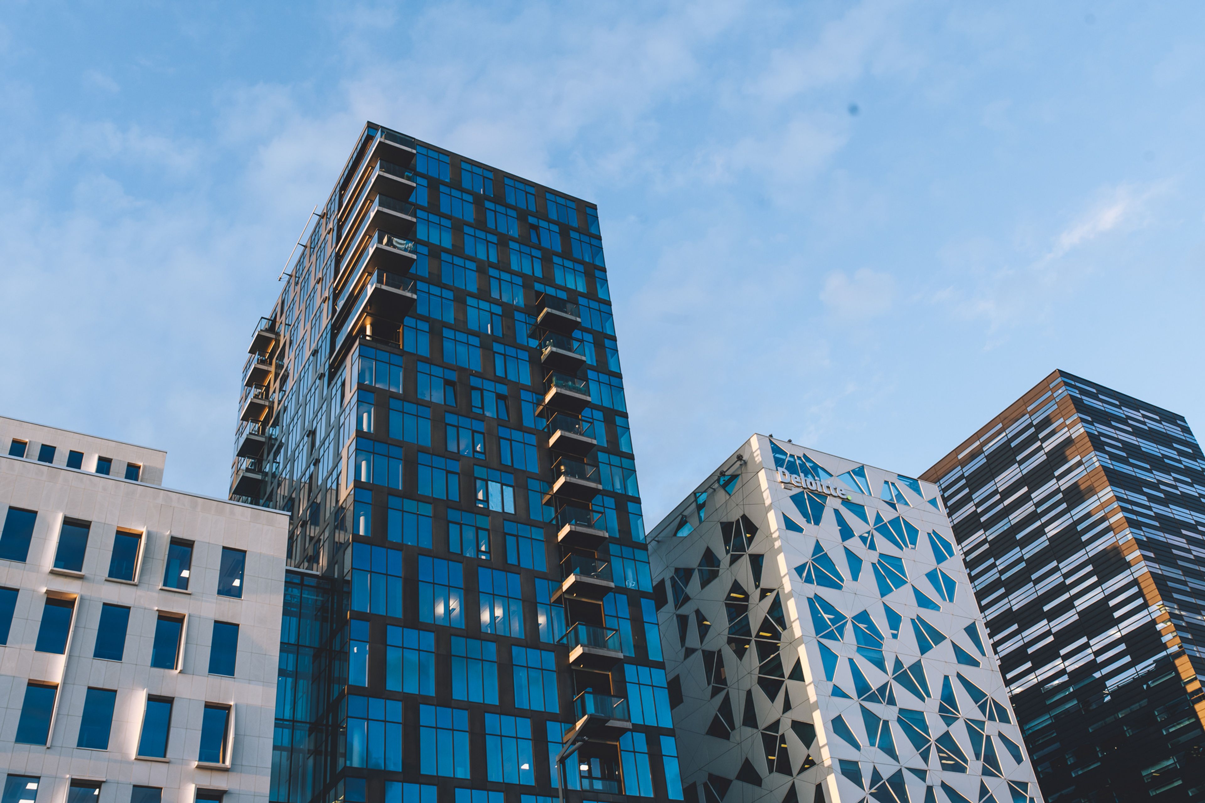 Utsikt nedenfra mot et høyt tårn med blå glassfasade ved siden av en hvit bygning med trekantmønstret fasade, mot en klar blå himmel.