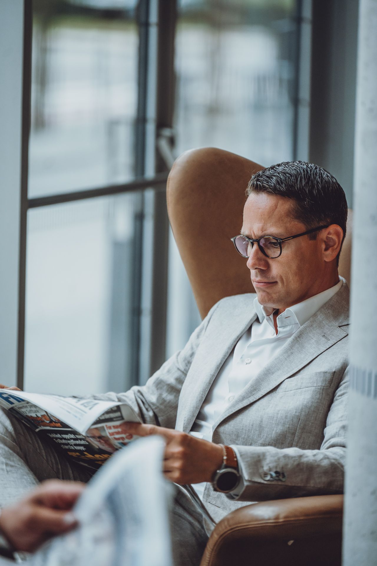 En mann i lys grå dress og briller sitter i en skinnlenestol og leser en finansavis ved et stort vindu.