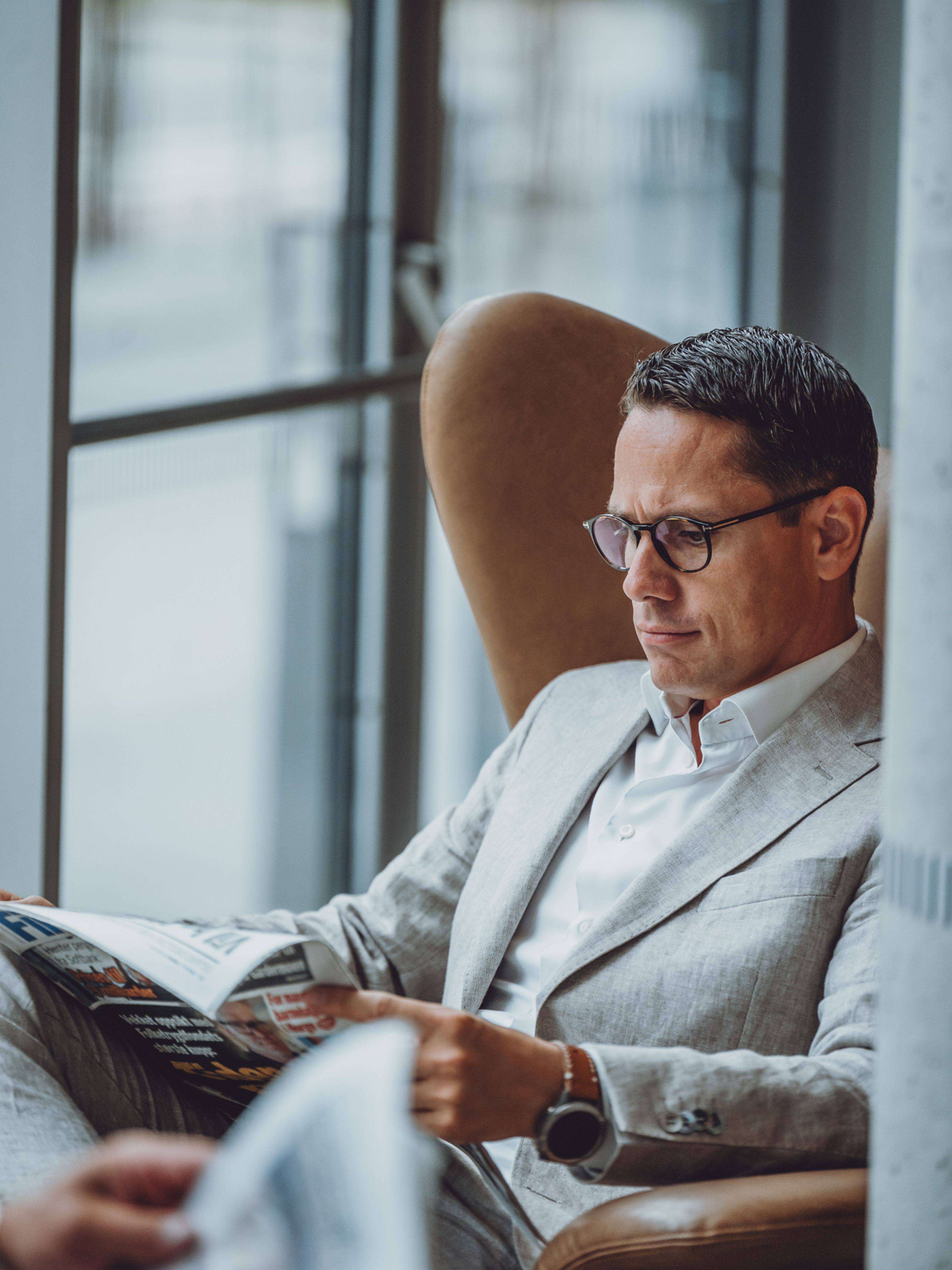 En mann i lys grå dress og briller sitter i en skinnlenestol og leser en finansavis ved et stort vindu.