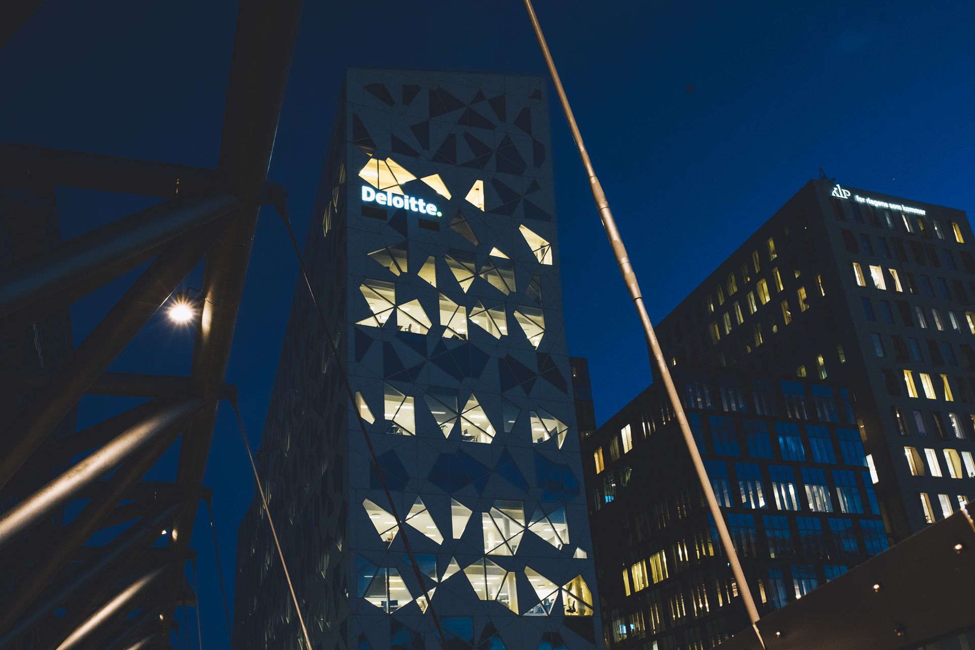 Nærbilde nedenfra av det samme opplyste kontorbygget med trekantet fasade og Deloitte-skilt om natten, med KLP-bygningen synlig ved siden av.
