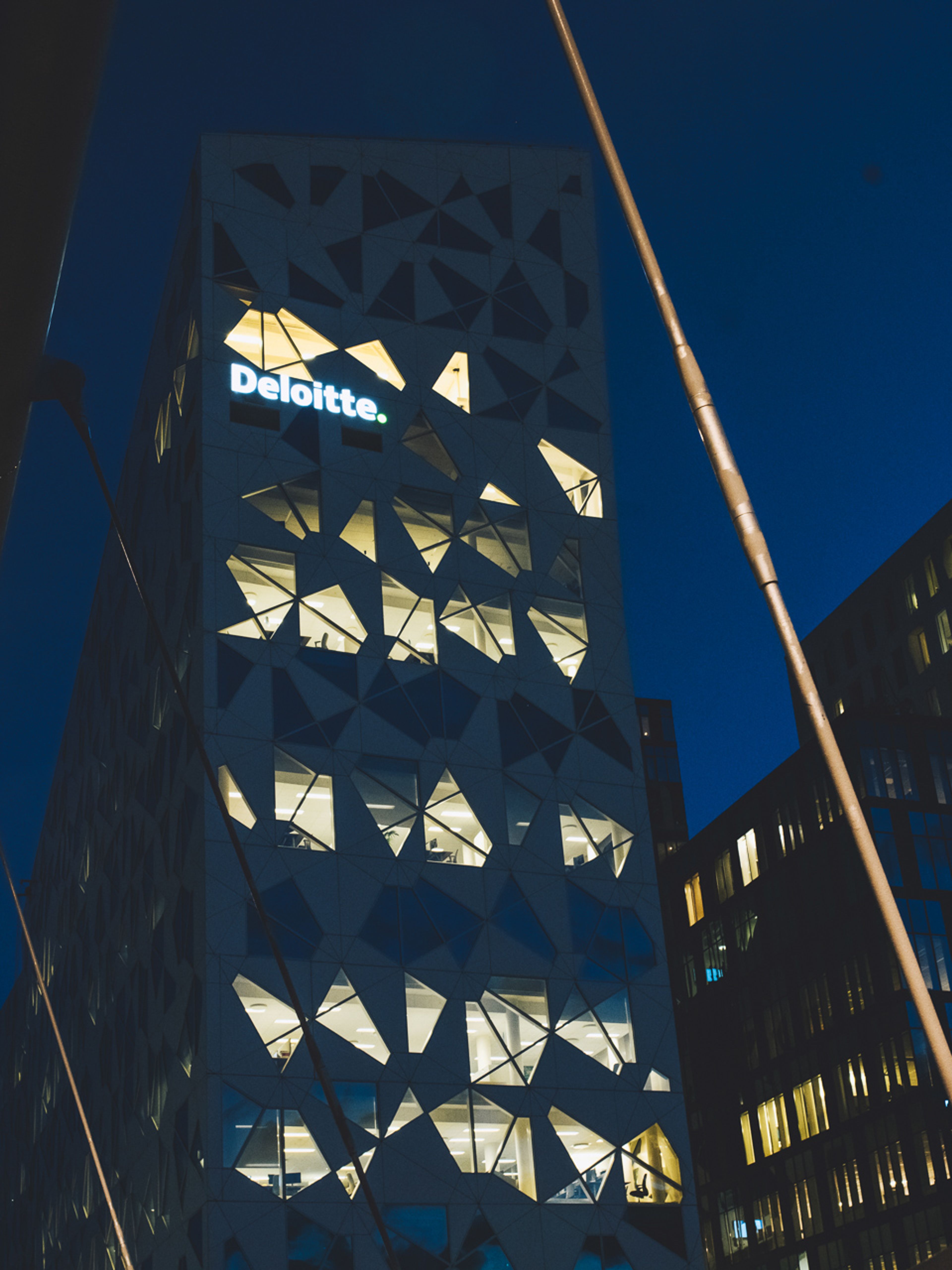 Nærbilde nedenfra av det samme opplyste kontorbygget med trekantet fasade og Deloitte-skilt om natten, med KLP-bygningen synlig ved siden av.