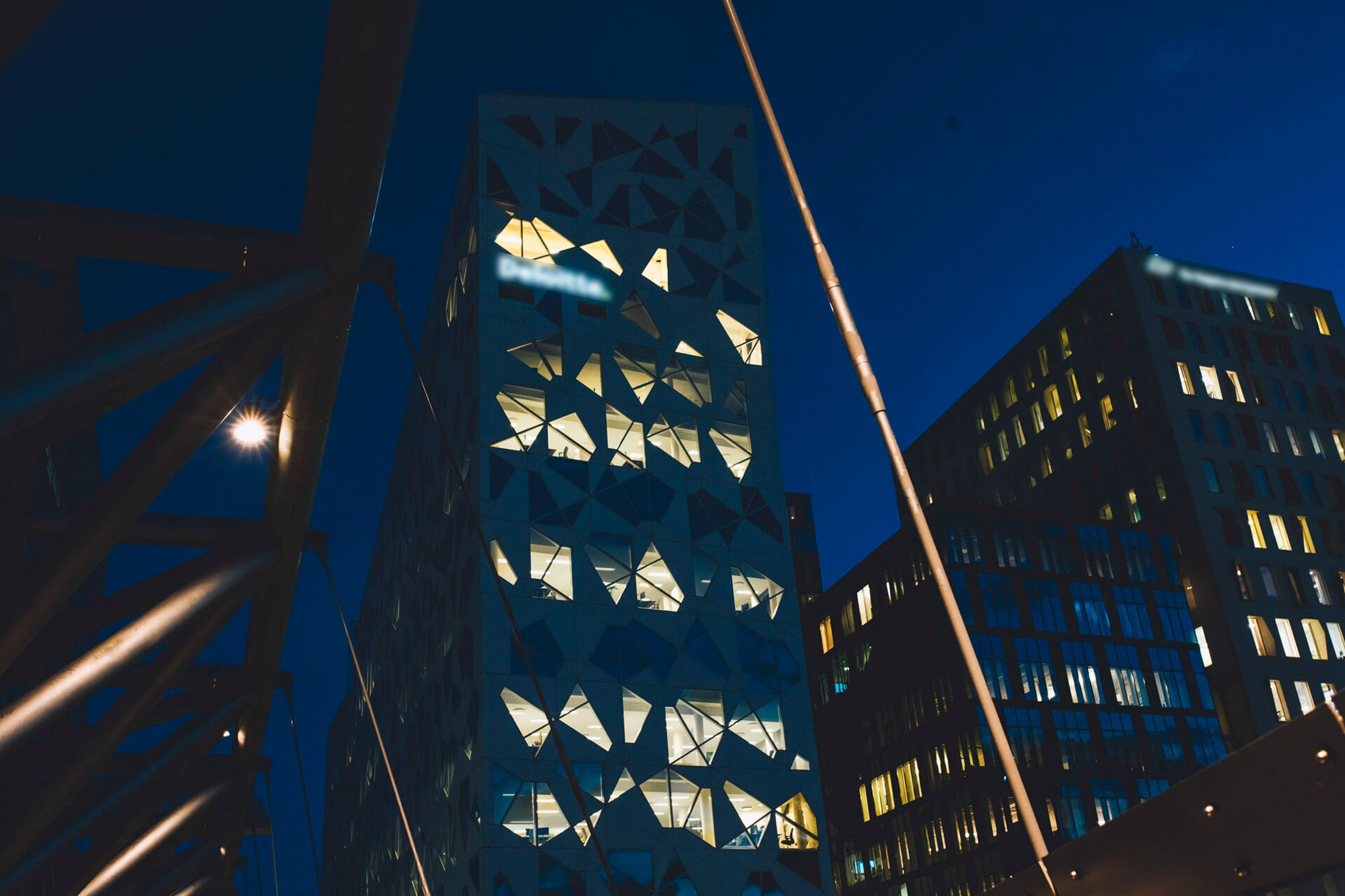 Det opplyste Deloitte-bygget i Bjørvika i Oslo i skumringen, sett fra en gangbro i stål.
