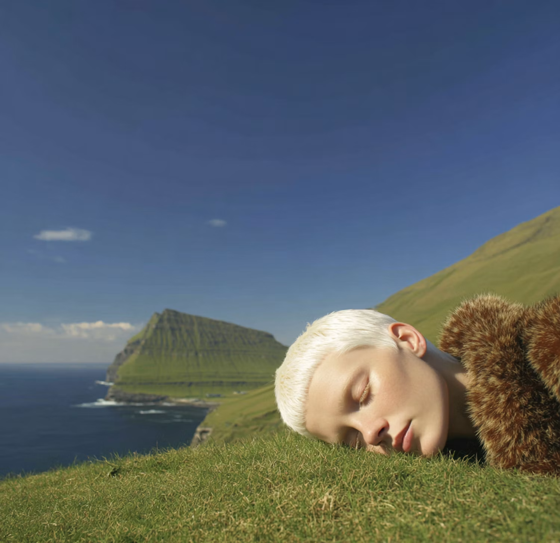 Person with white hair lying on a grassy cliff by the sea