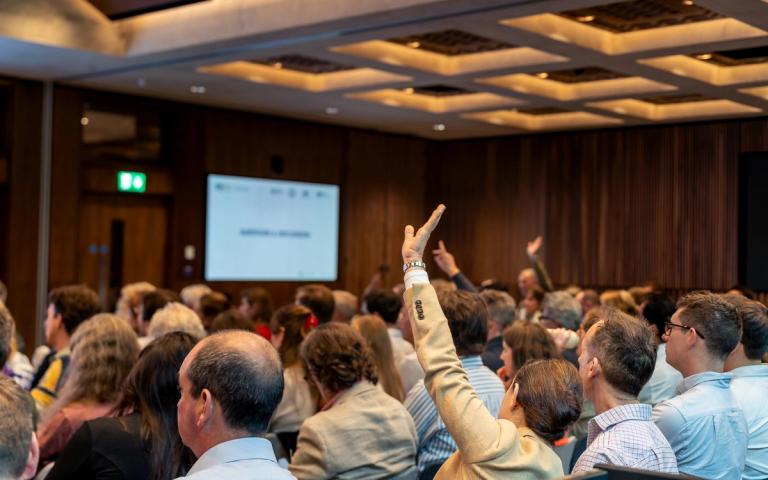Audience at a conference with several hands raised for questions.