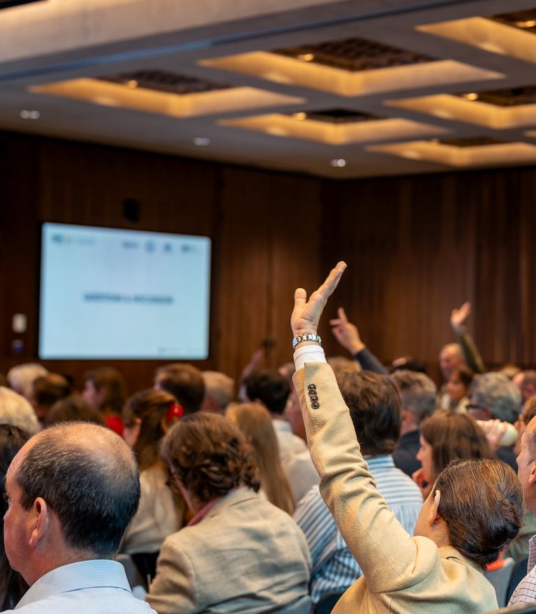 Audience at a conference with several hands raised for questions.