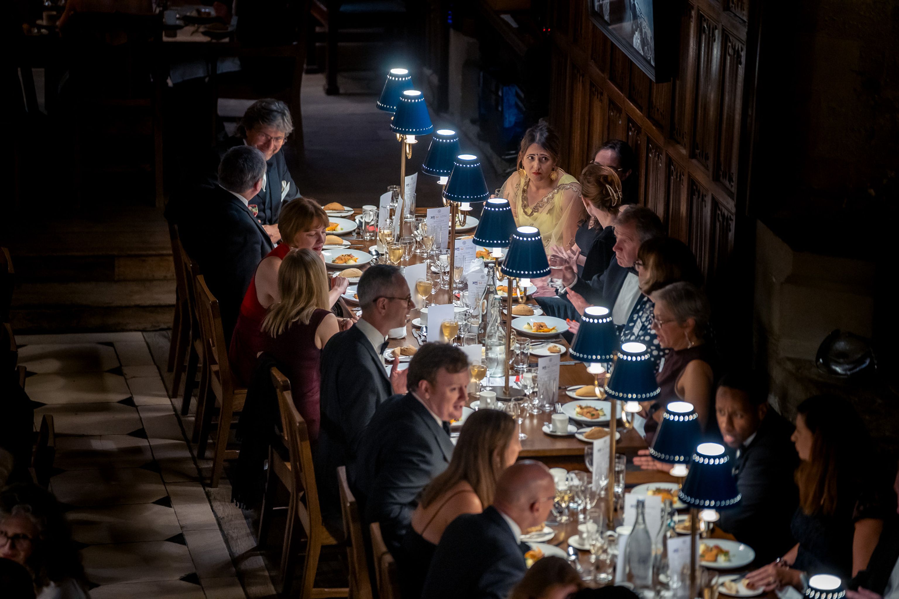 Guests in formal attire seated at a long table under blue-shaded lamps in a dimly lit hall.