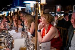 A woman in a red dress sits attentively at a long banquet table under blue-shaded lamps.