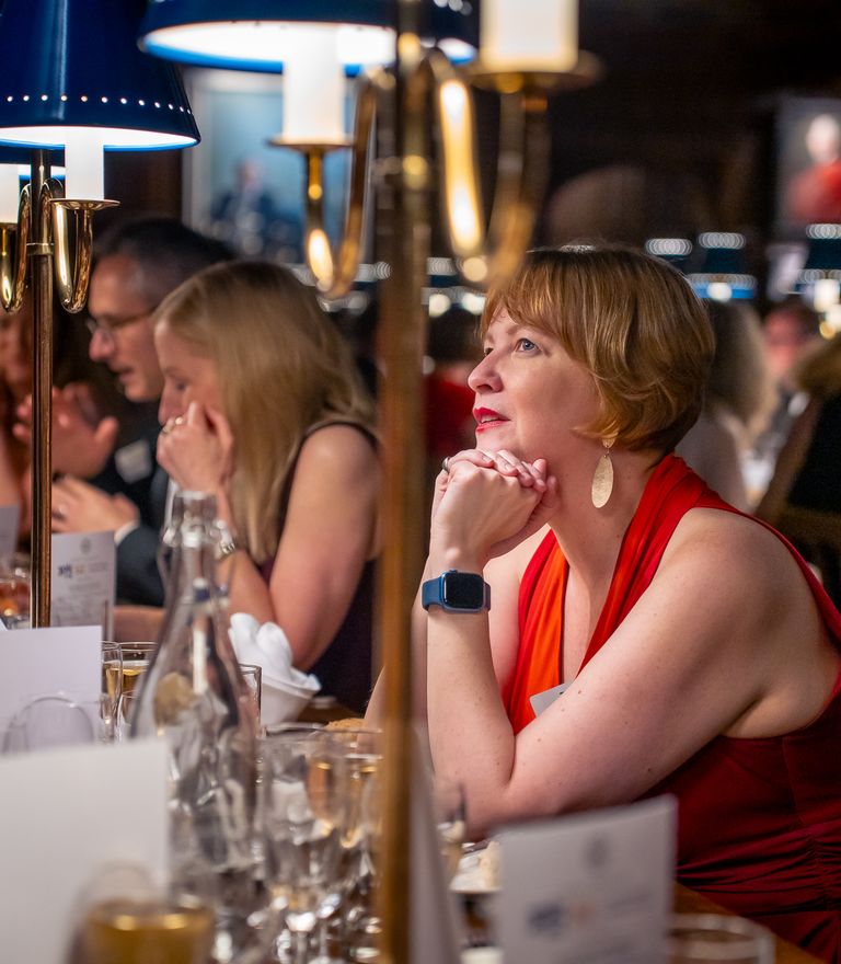 A woman in a red dress sits attentively at a long banquet table under blue-shaded lamps.