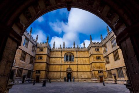 View through an arched entryway into a historic stone quadrangle building with Gothic architecture under a blue sky.