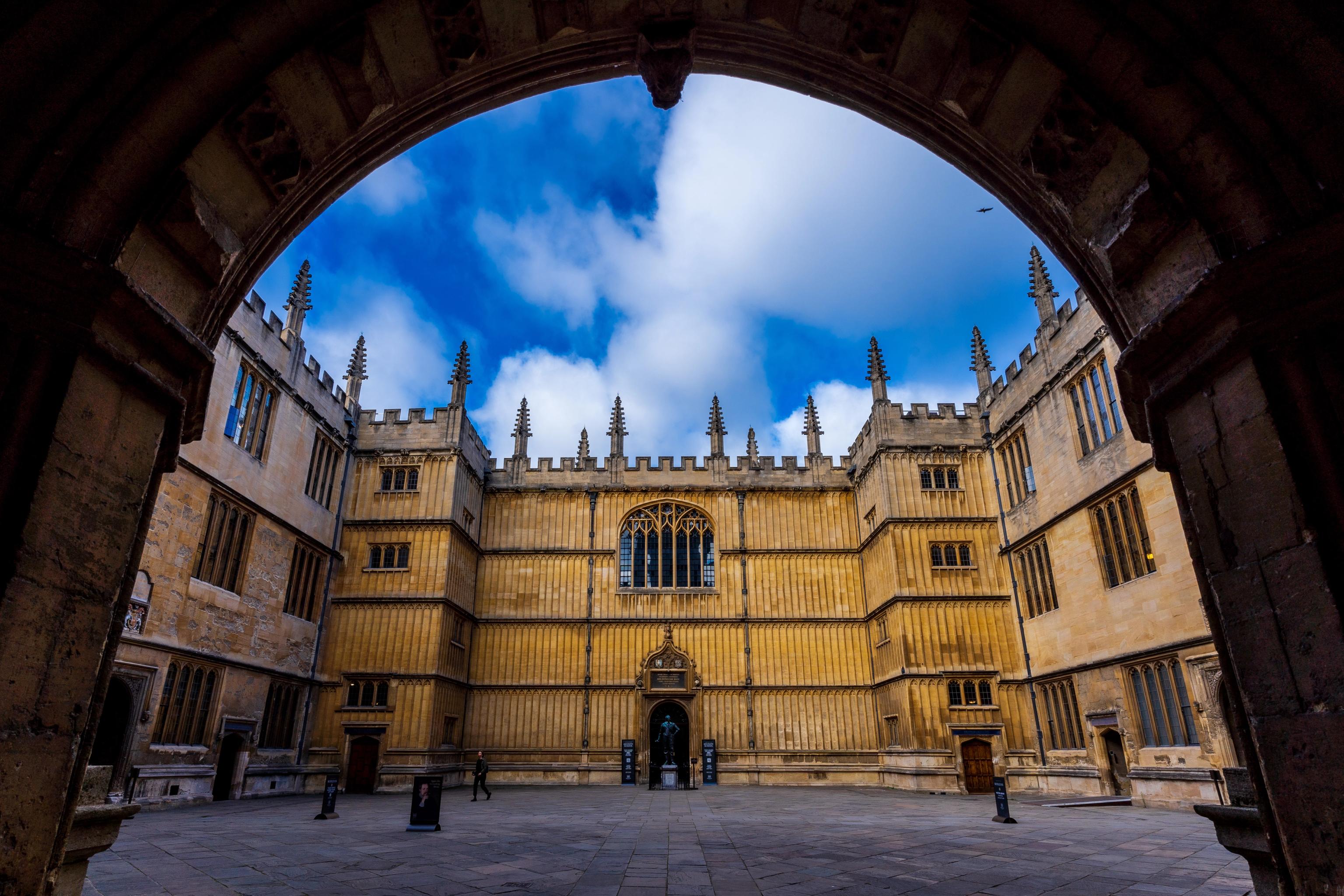 View through an arched entryway into a historic stone quadrangle building with Gothic architecture under a blue sky.
