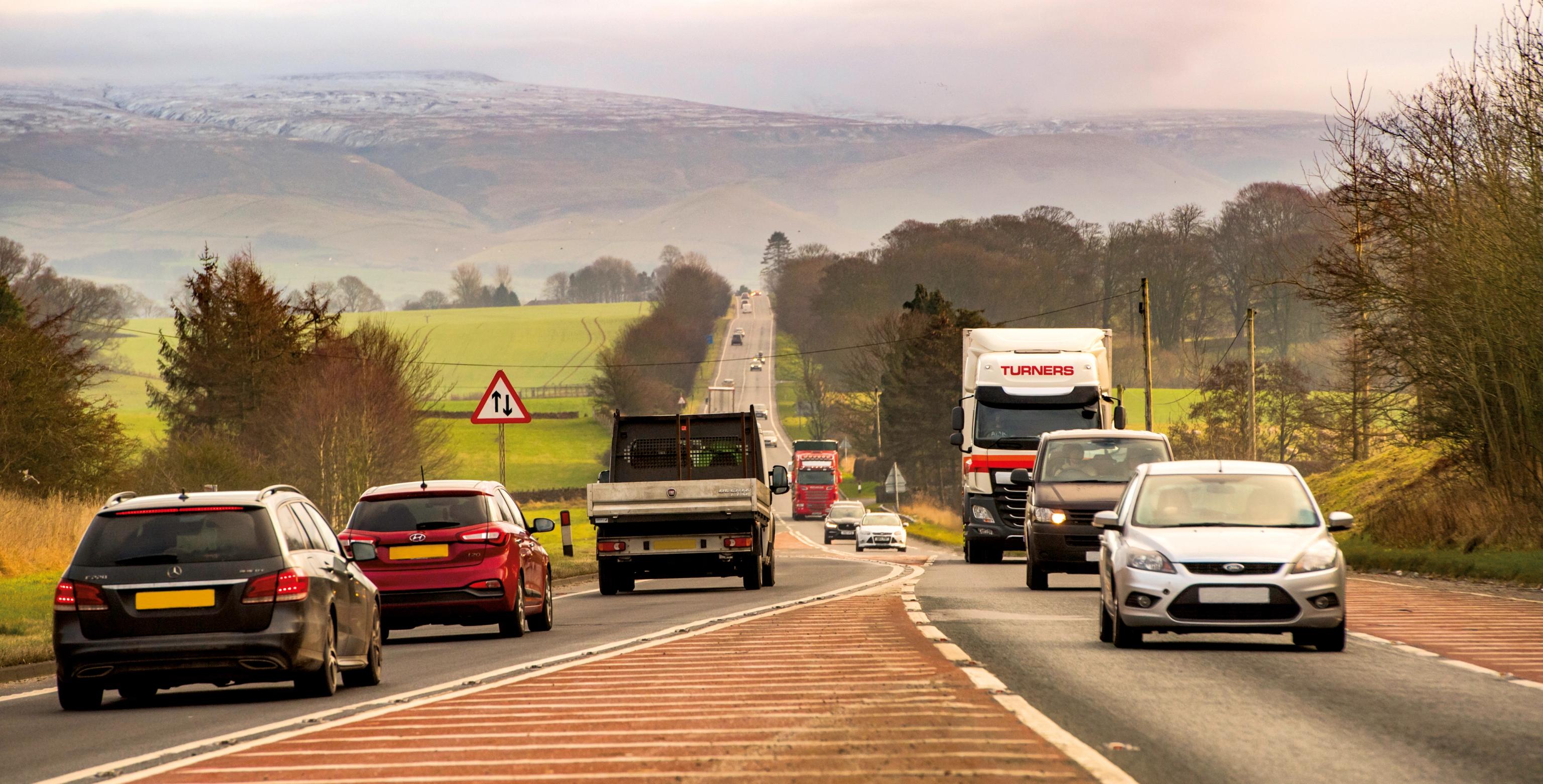 a row of cars are driving down a country road .
