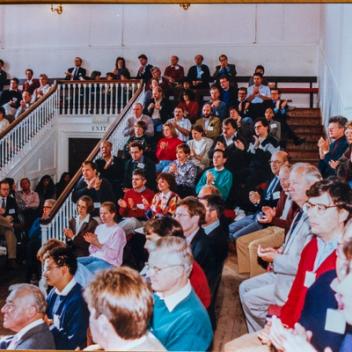 A large audience fills an auditorium with multi-level seating, many people are clapping.