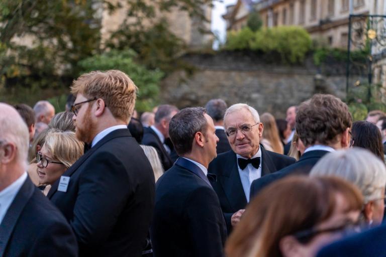 A crowd of formally dressed people socialize outdoors, with an older man in a tuxedo smiling at the viewer.