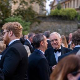 A crowd of formally dressed people socialize outdoors, with an older man in a tuxedo smiling at the viewer.
