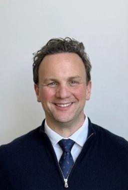 Headshot of a smiling man with dark wavy hair wearing a tie and a navy quarter-zip sweater.