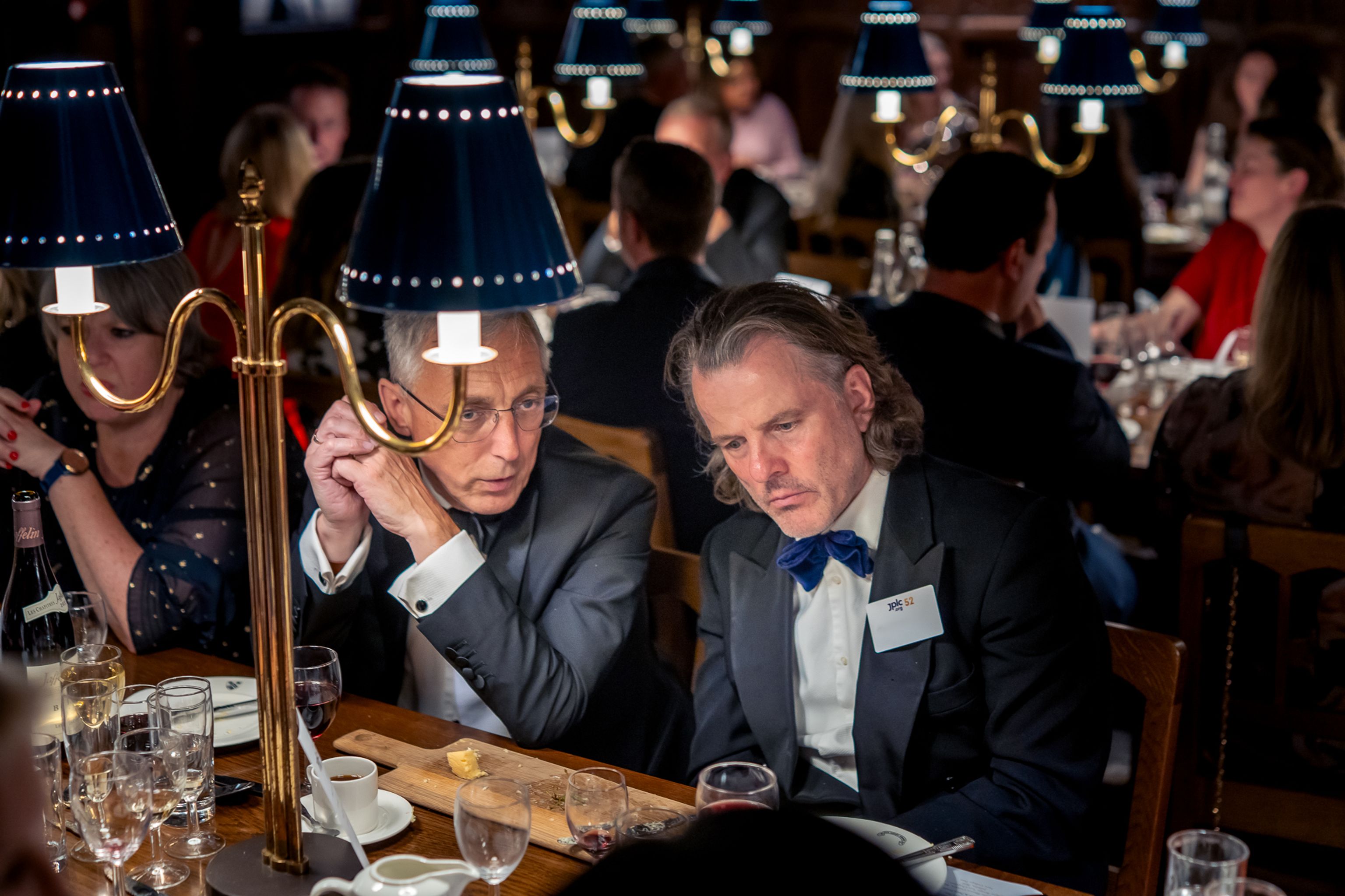 Two men in formal wear seated at a table with a lamp during a dinner event.