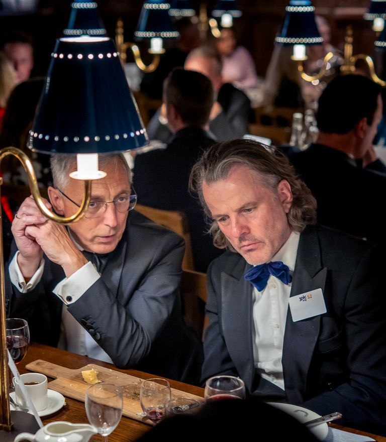 Two men in formal wear seated at a table with a lamp during a dinner event.