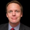 A smiling man with light brown hair wears a pinstripe suit and red tie.