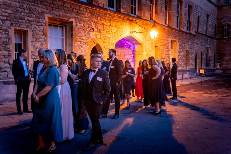A group of well-dressed people socialize outdoors at night in a stone courtyard, illuminated by warm orange light and a purple-lit archway.