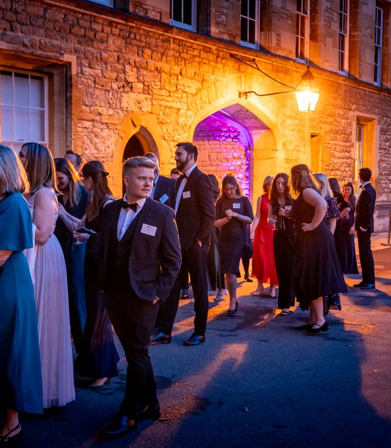 A group of well-dressed people socialize outdoors at night in a stone courtyard, illuminated by warm orange light and a purple-lit archway.