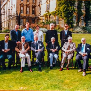 A group of thirteen well-dressed people, mostly men, pose on a sunny green lawn in front of a historic building.