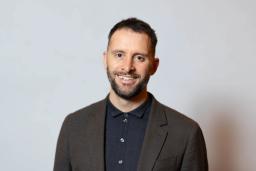 A smiling man with short dark hair and a beard, wearing a dark jacket and polo shirt, against a light background.