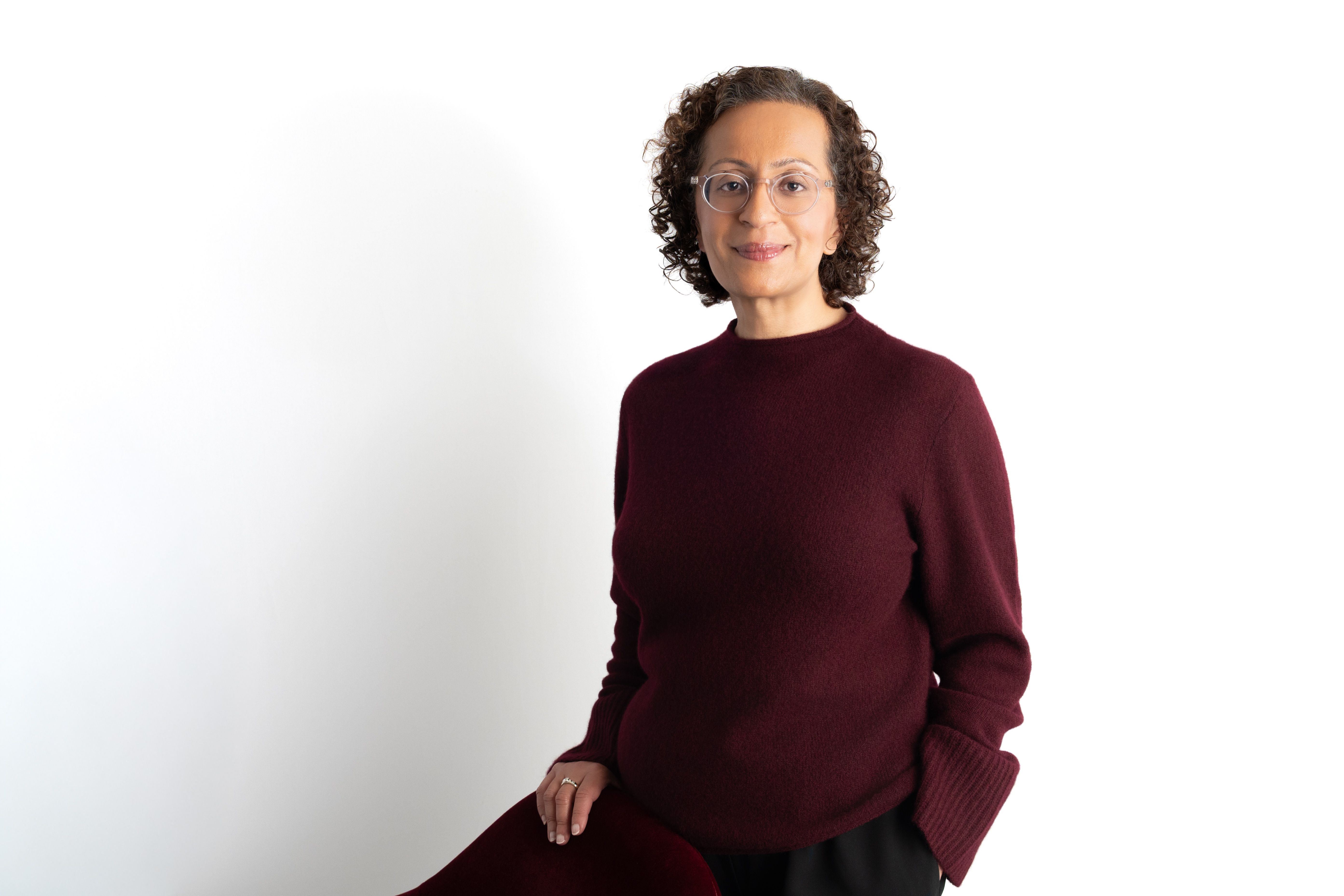 A woman with curly hair and glasses, wearing a burgundy sweater, smiles gently against a white background.