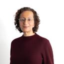 A woman with curly hair and glasses, wearing a burgundy sweater, smiles gently against a white background.