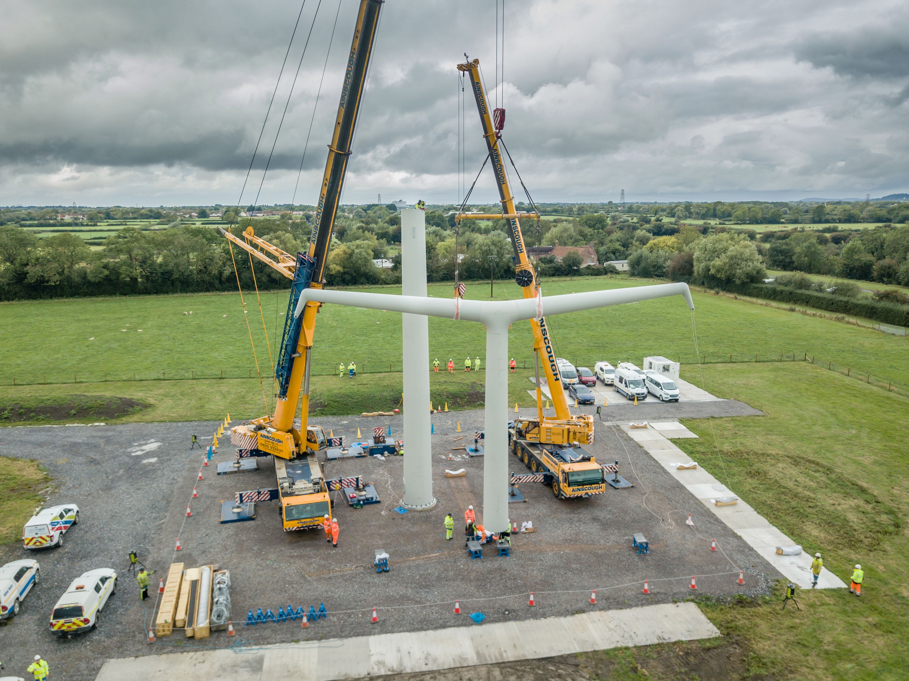 Two cranes lifting a large Y-shaped wind turbine rotor next to its tower on a construction site.