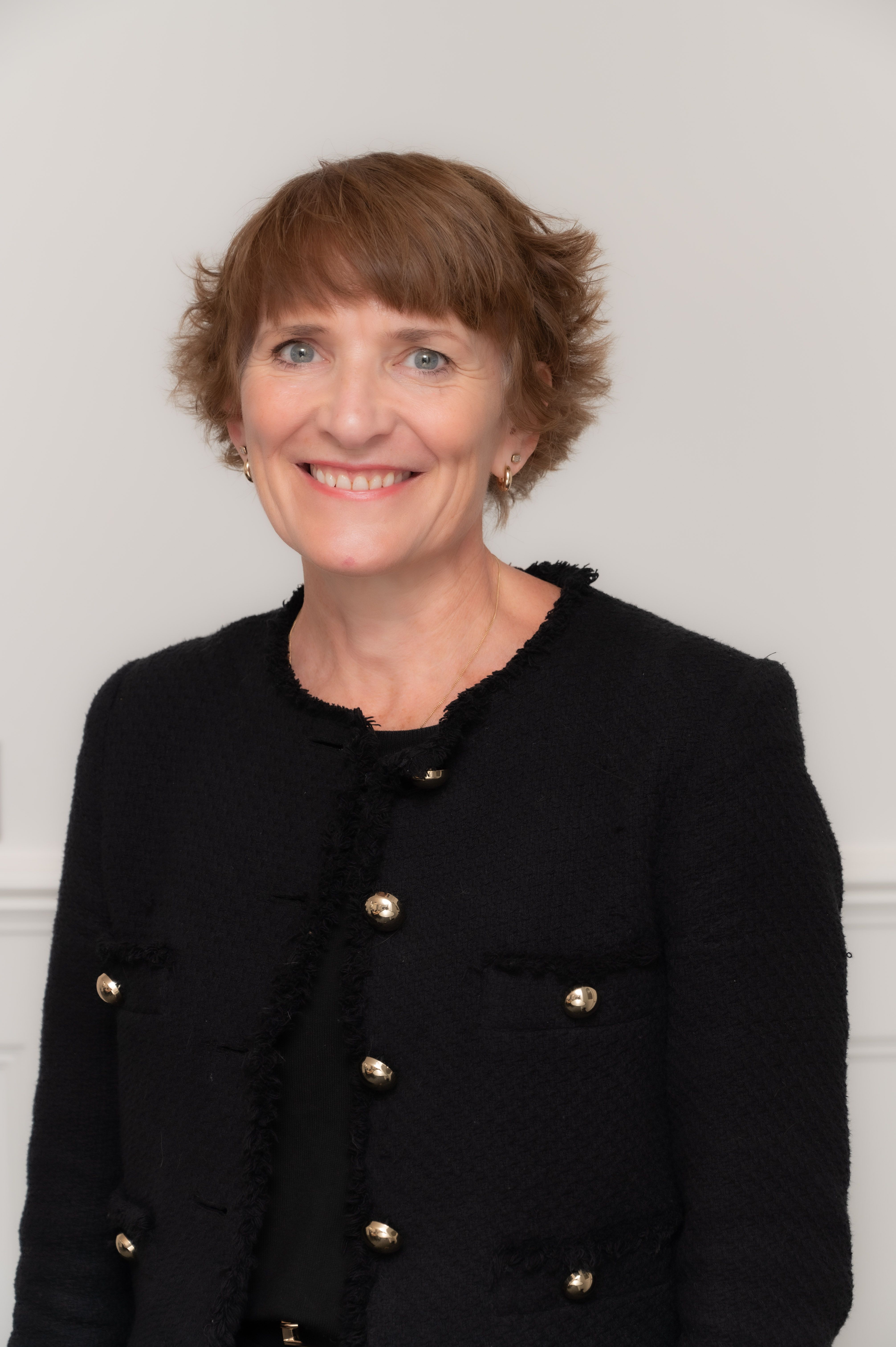 A smiling woman with short, reddish-brown hair and blue eyes wears a black tweed jacket with gold buttons.