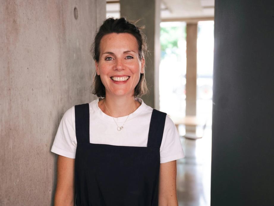 A smiling woman with long brown hair, glasses, a blue top, and yellow earrings, with arms crossed against a brick wall.