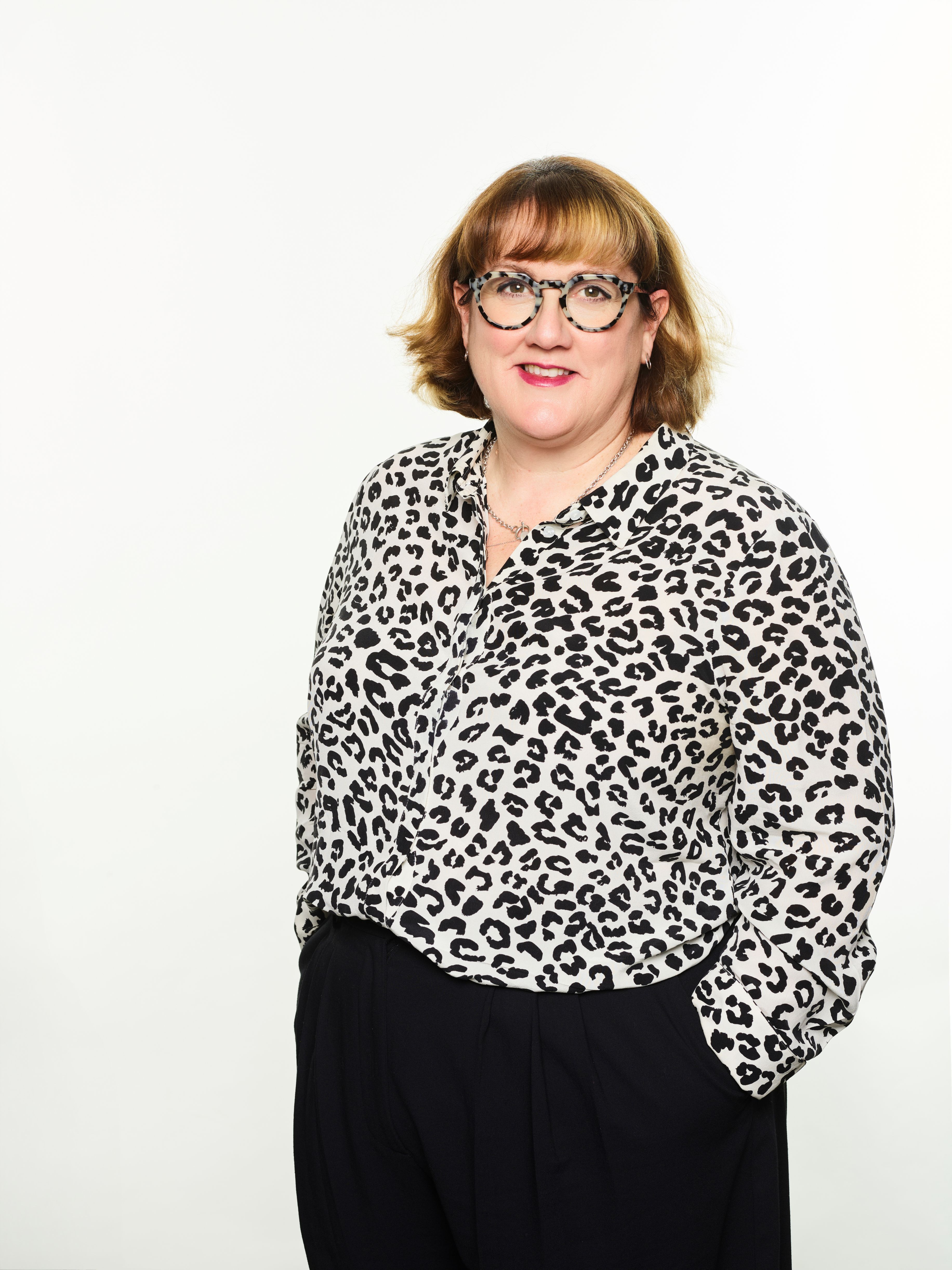 Smiling woman in a leopard print shirt, black pants, and glasses, standing with hands in pockets.