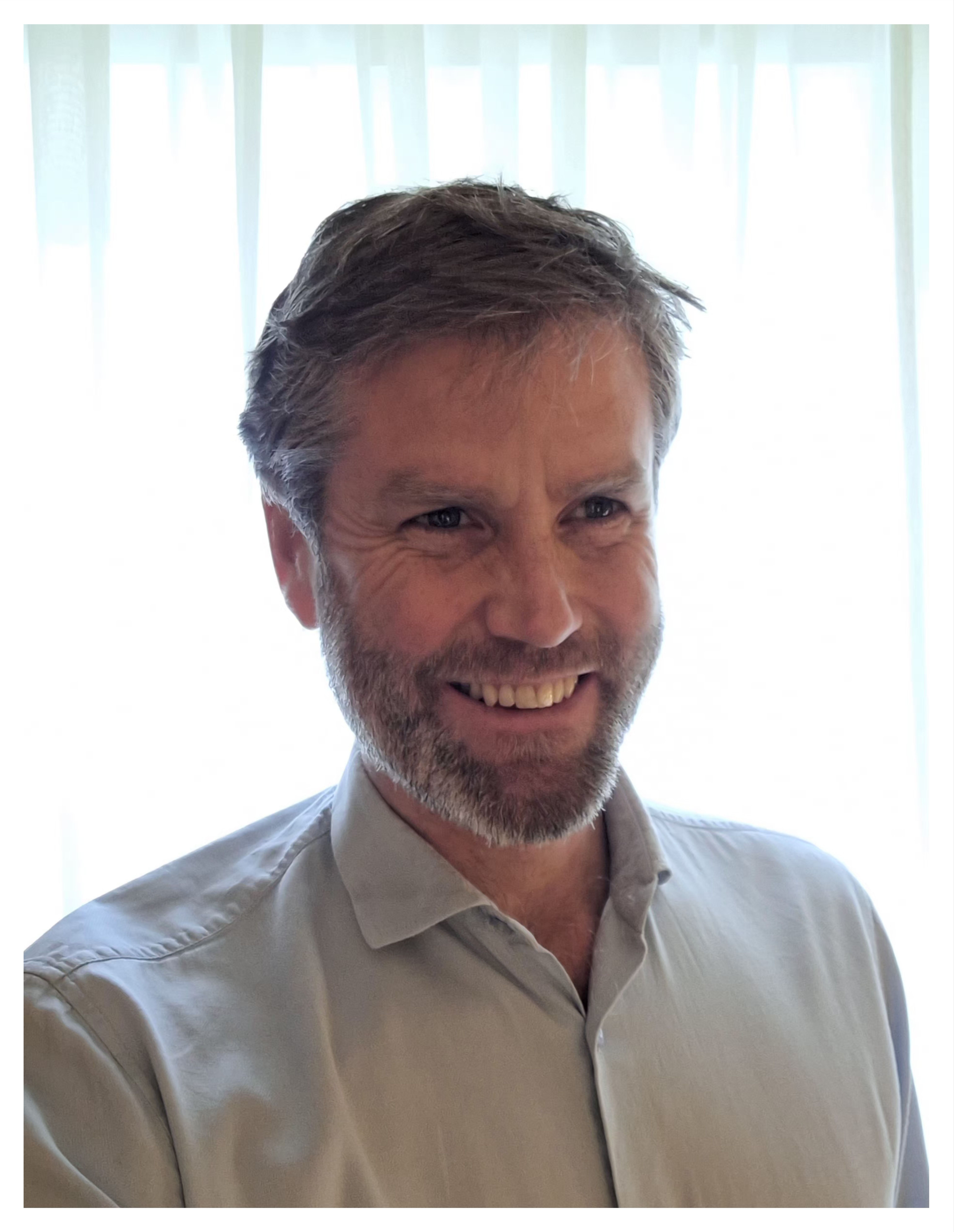 Smiling man with grey hair and a beard, wearing a light shirt.