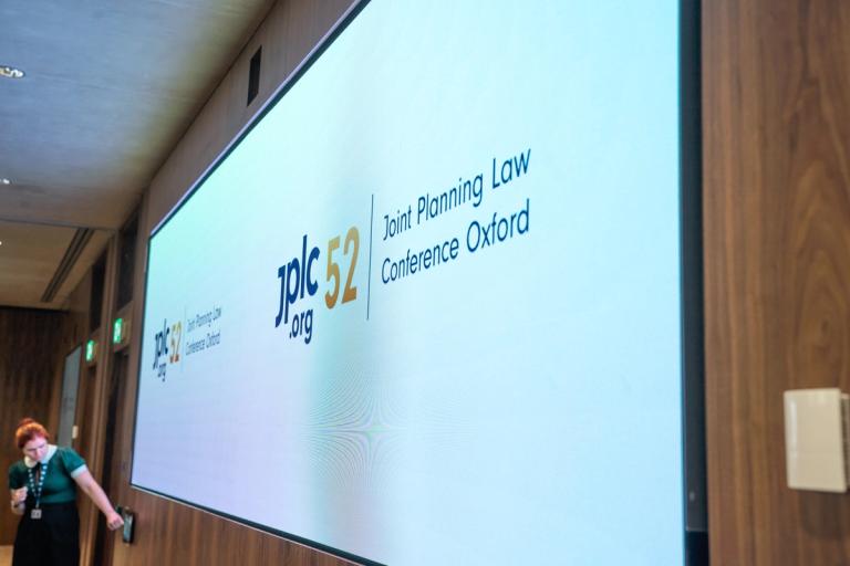 A large screen displays "jplc 52 .org" and "Joint Planning Law Conference Oxford" in a wood-paneled room with a person standing to the left.