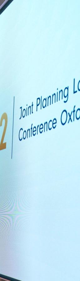 A large screen displays "jplc 52 .org" and "Joint Planning Law Conference Oxford" in a wood-paneled room with a person standing to the left.