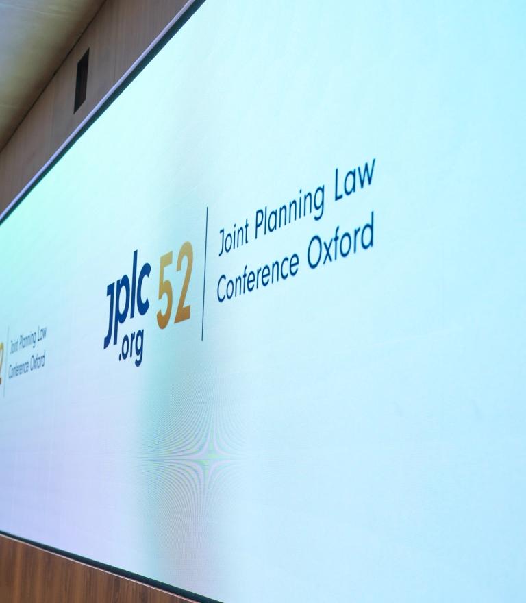 A large screen displays "jplc 52 .org" and "Joint Planning Law Conference Oxford" in a wood-paneled room with a person standing to the left.