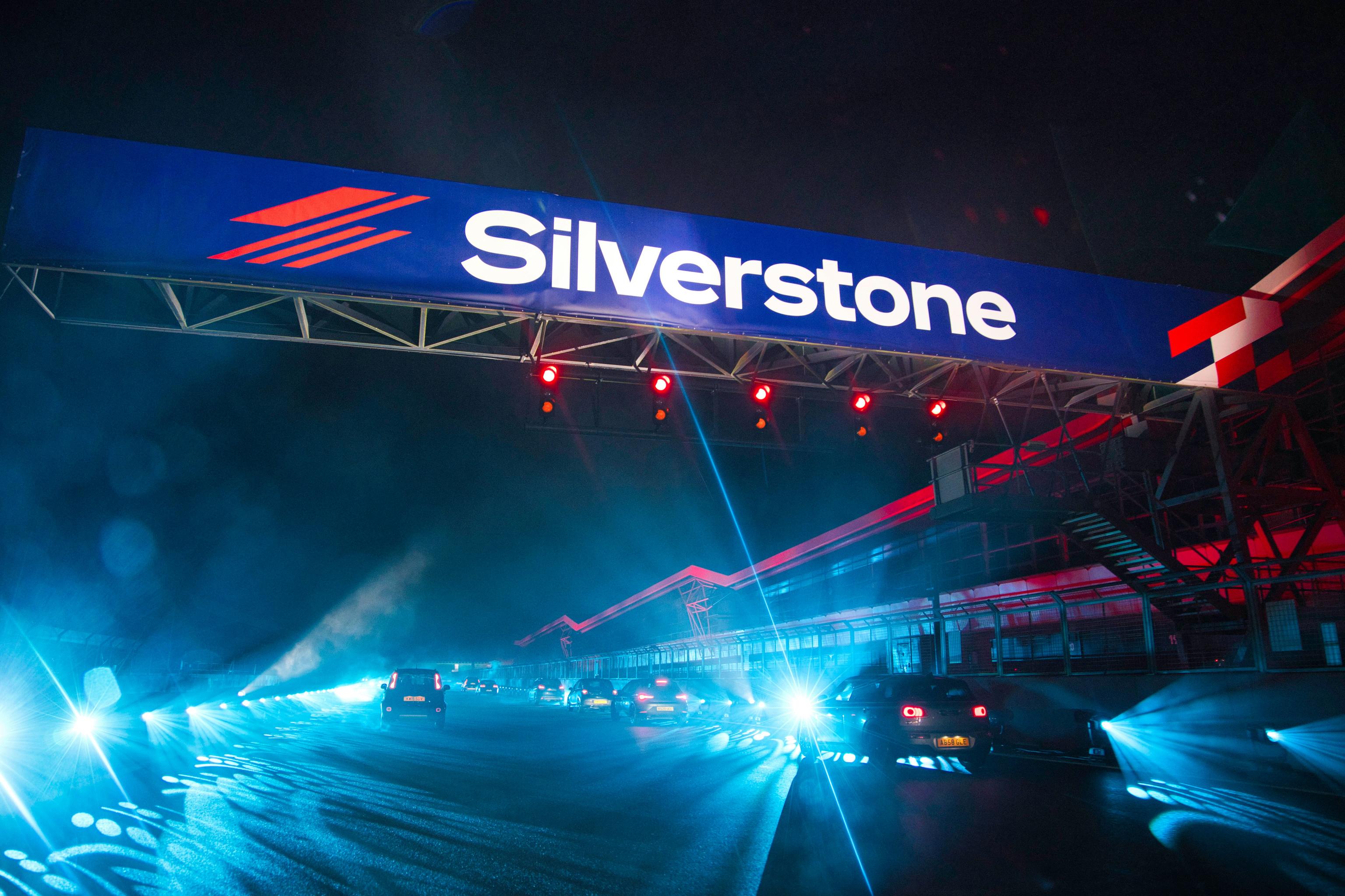 Night view of Silverstone racetrack with cars illuminated by bright blue lights under a large Silverstone sign.