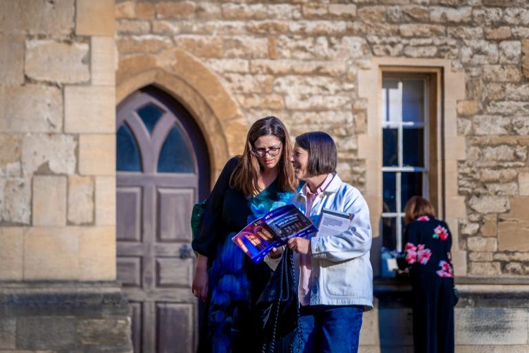 Two women look at a brochure in front of a stone building.