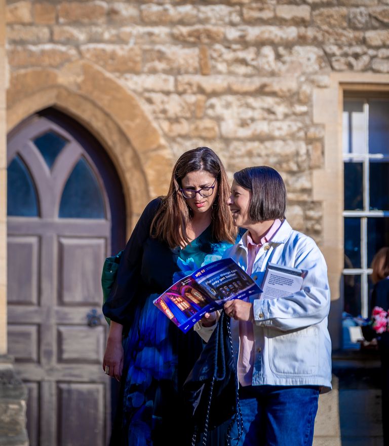 Two women look at a brochure in front of a stone building.