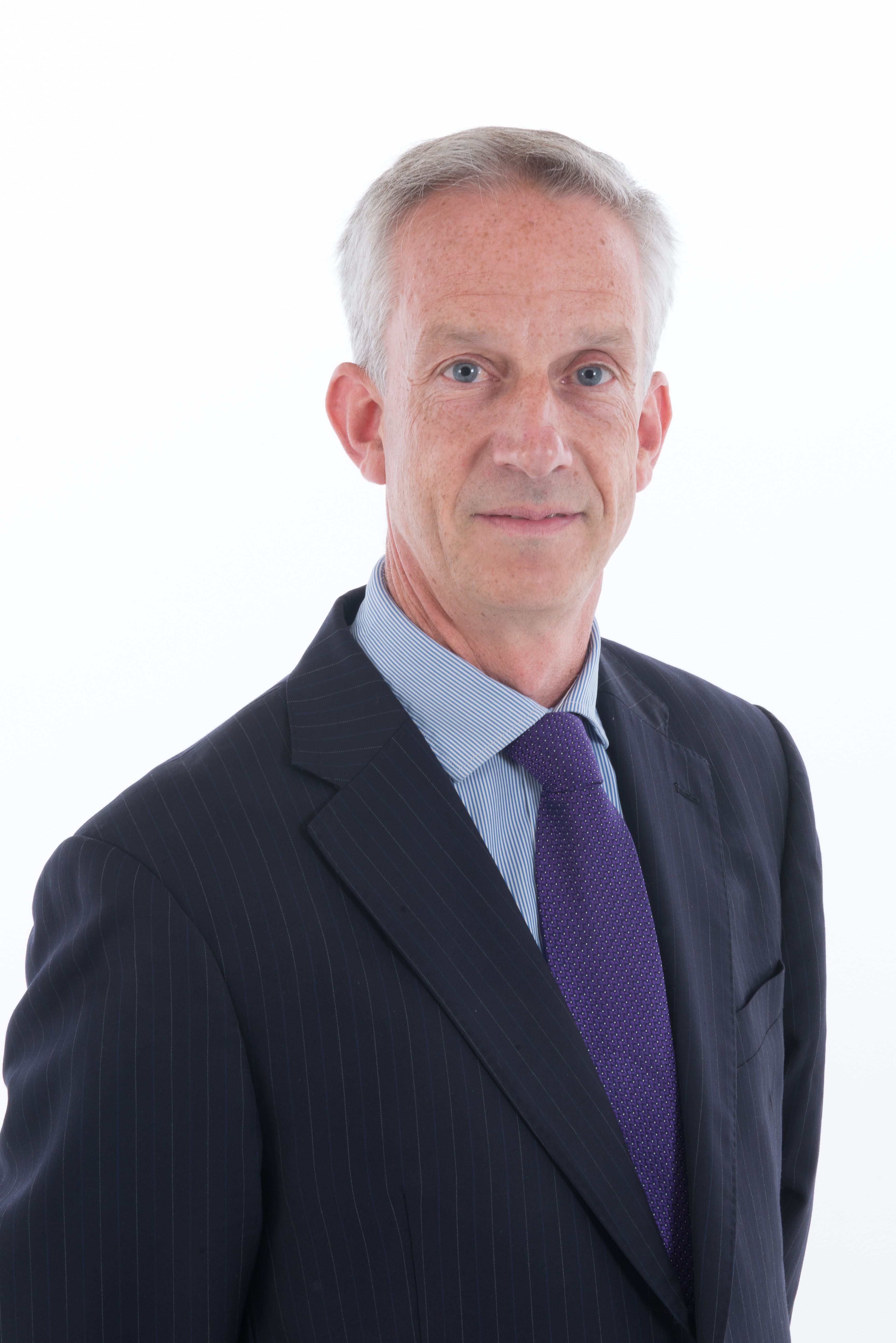 A man with grey hair and blue eyes, wearing a dark pinstripe suit and purple tie.