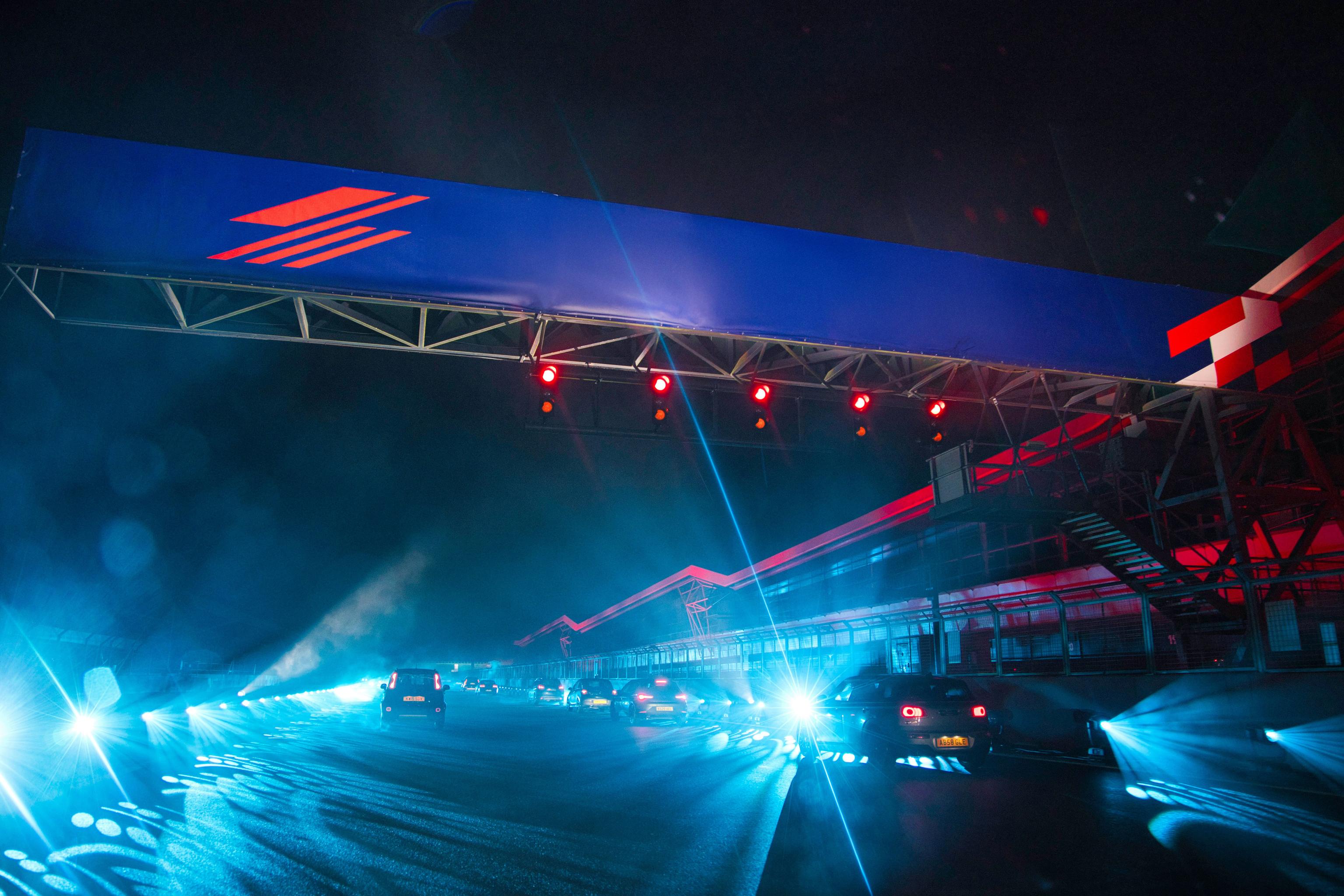 Night view of Silverstone racetrack with cars illuminated by bright blue lights under a large Silverstone sign.