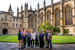 A diverse group of twelve people stands in the courtyard of a historic stone building with a green lawn.