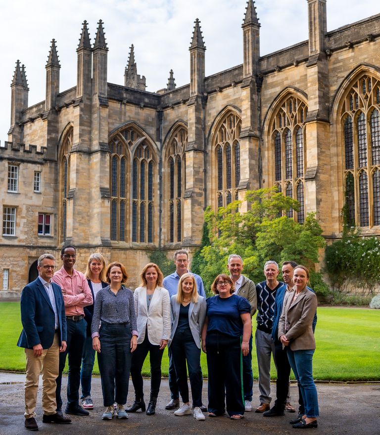 A diverse group of twelve people stands in the courtyard of a historic stone building with a green lawn.