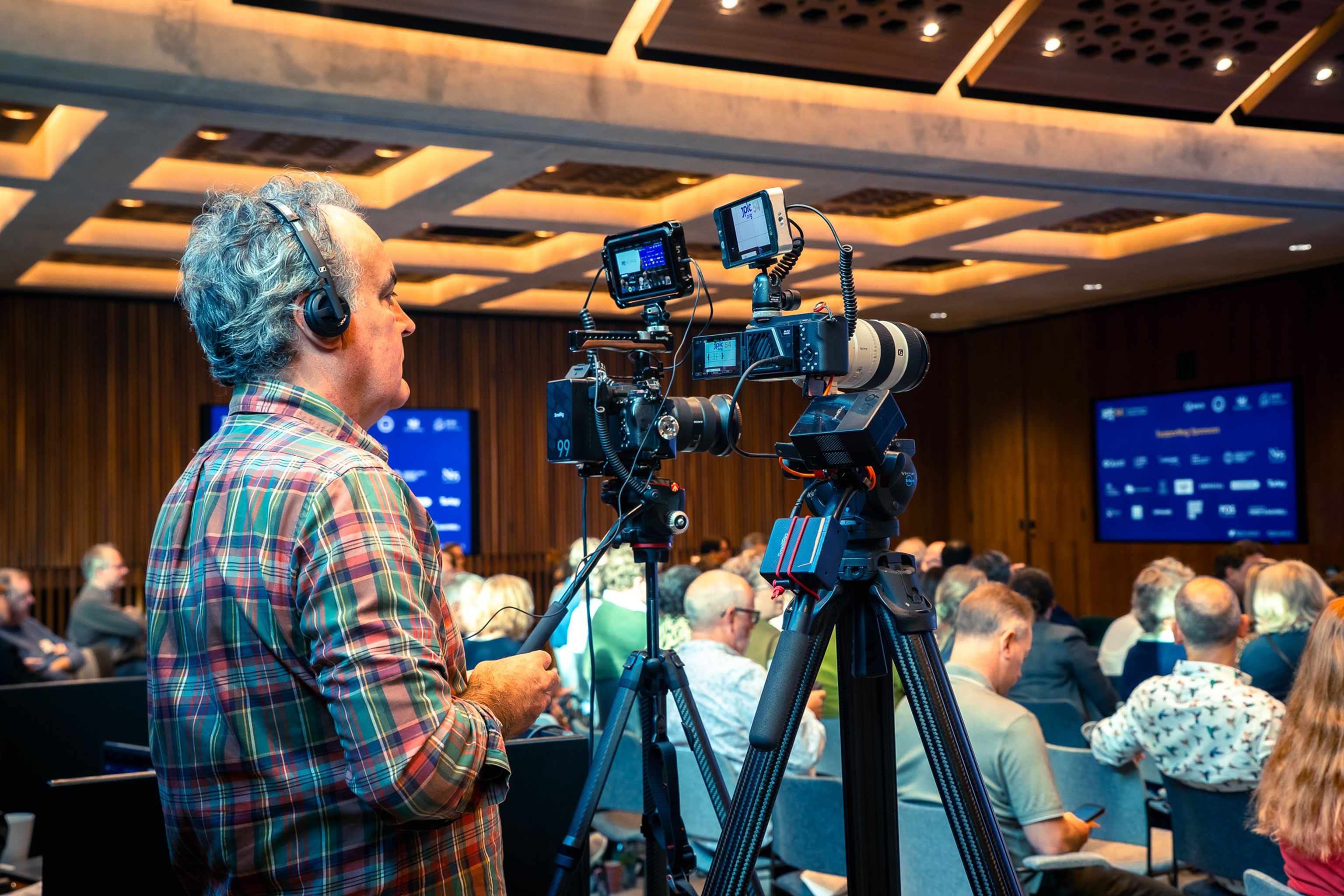 An audience seated in a conference room, watching two speakers on a stage with a screen displaying "Opening Address."