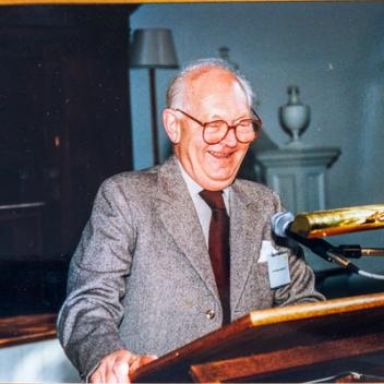 A man in a suit and glasses laughs heartily behind a podium with a microphone.