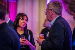 A woman and a man chat, holding drinks, at a social event.